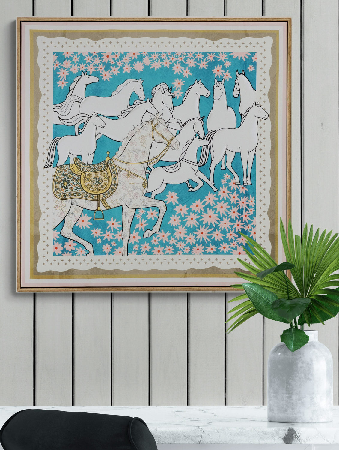 Framed artwork with horses and floral patterns on a white wall with a plant.
